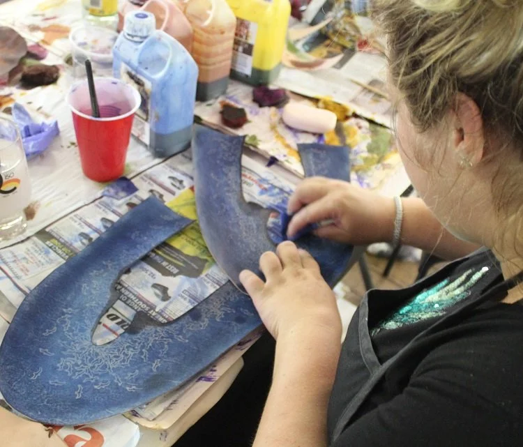 A woman is painting a blue shoe with intricate designs at a  table covered with paint bottles, and supplies.
