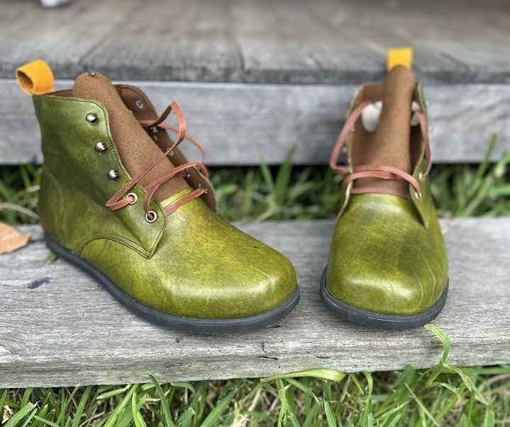 Pair of olive green leather boots with brown accents and pink laces placed on a wooden surface with grass underneath.