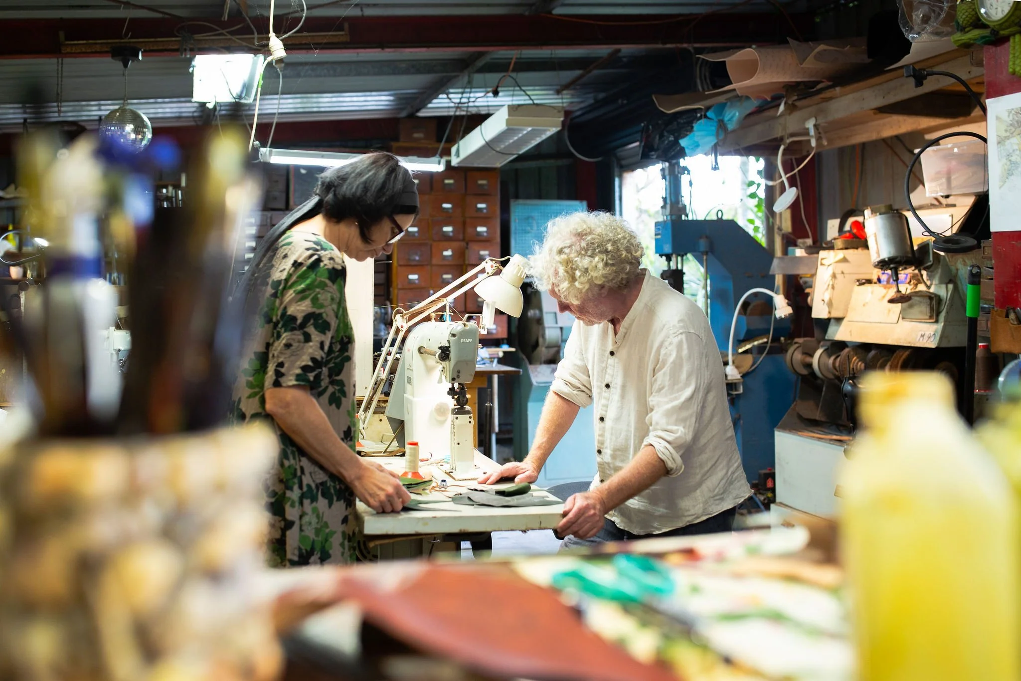 Two women are working together at a cluttered workbench in a workshop, focused on a project with fabric and a sewing machine, surrounded by various tools and supplies.