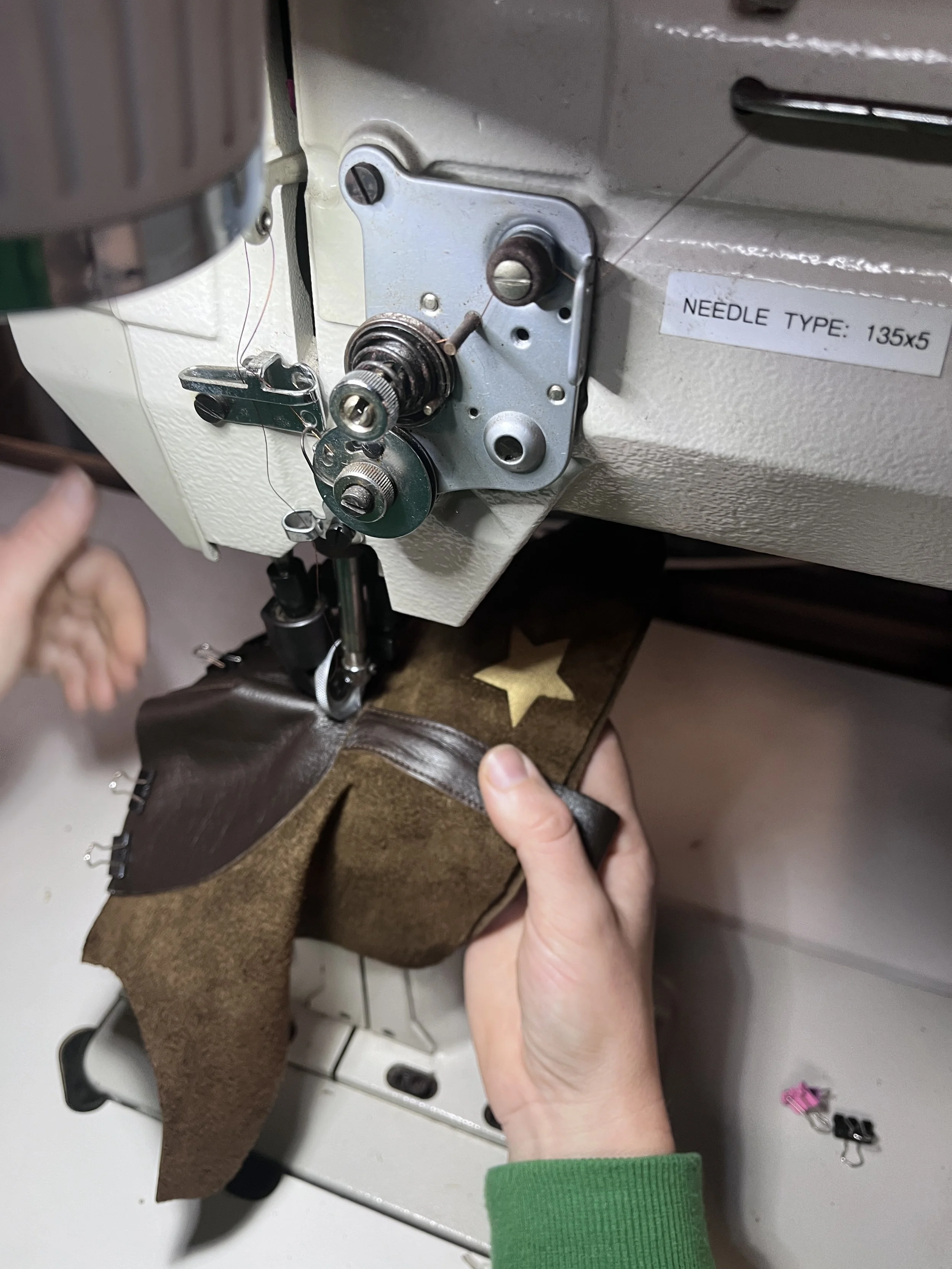 Close-up of a shoemakers' post bed sewing machine with a person's hands guiding a piece of brown leather  through it.