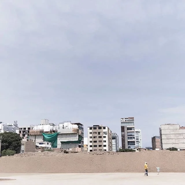 Cityscape with several buildings, some under construction, under a cloudy sky, with a person walking on a wide open paved area in the foreground.