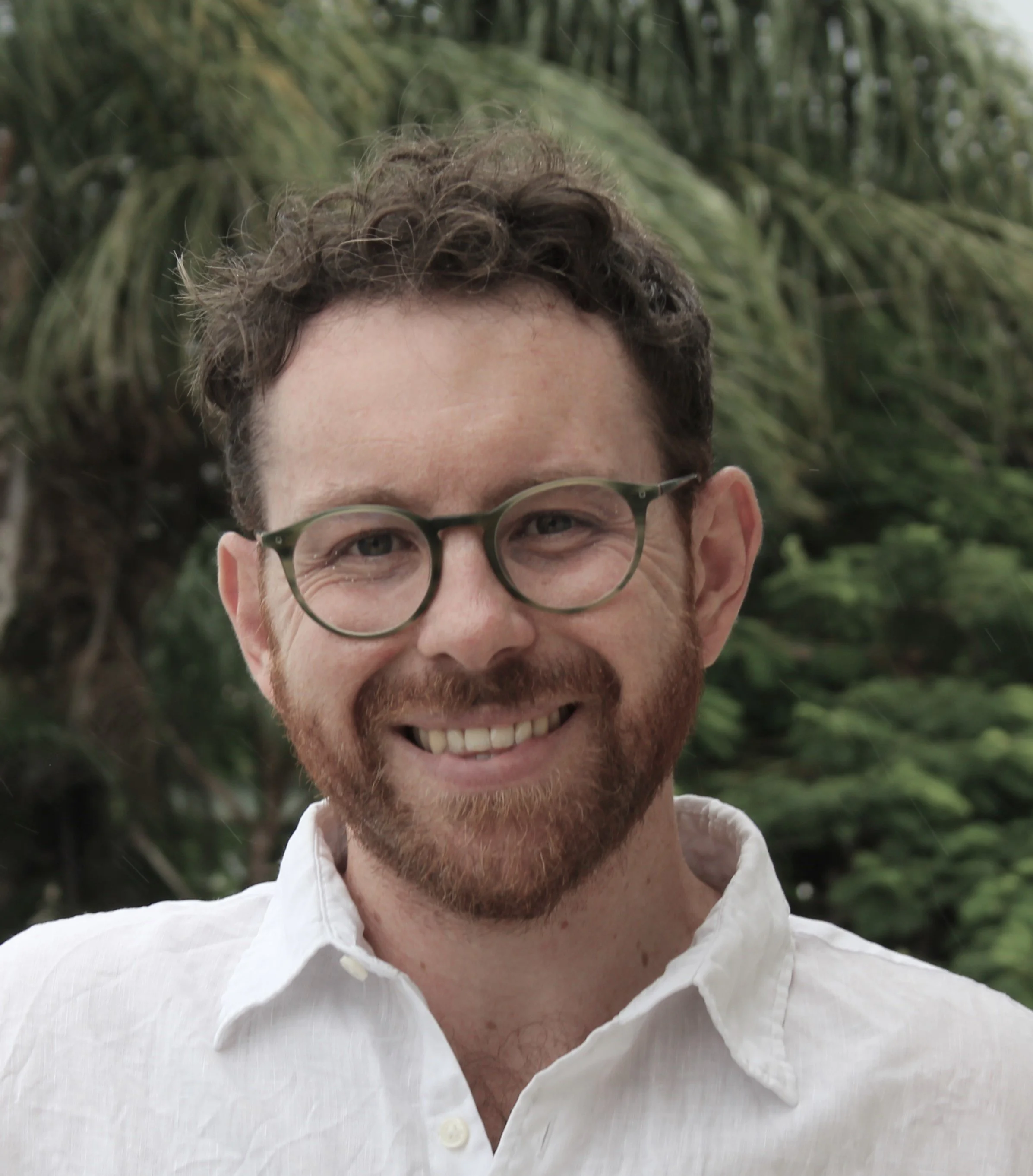 Close-up of a smiling man with curly brown hair, glasses, and a beard, wearing a white shirt, standing outdoors with green trees in the background.