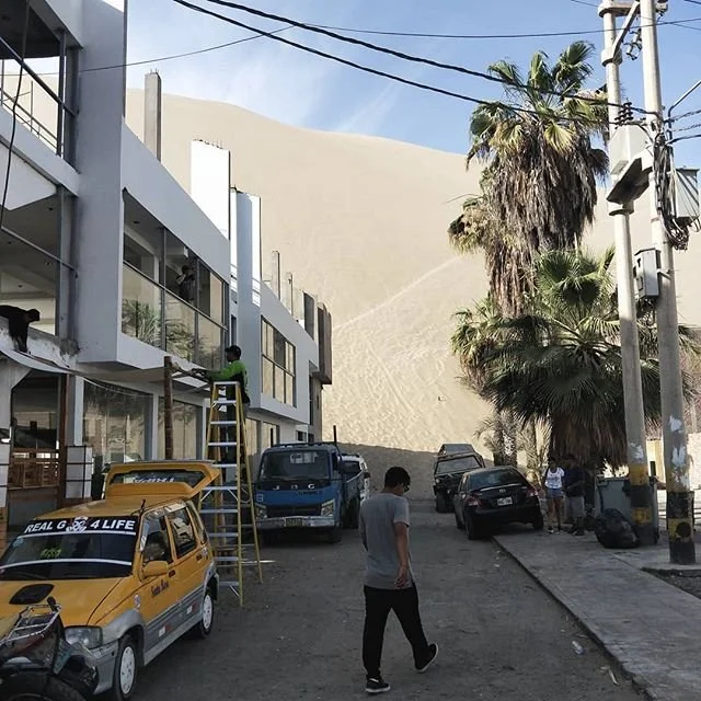 People stand near cars on a sandy street with a multi-story building, tall trees, and a large sand dune in the background.