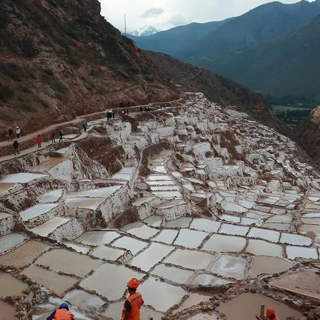 Terraced salt evaporation ponds on a mountainside with hikers walking along a trail above.