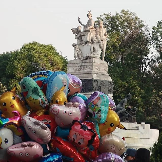 A monument with a classical sculpture of people and animals at the top, surrounded by trees, with a large pile of colorful balloons featuring cartoon characters in the foreground.