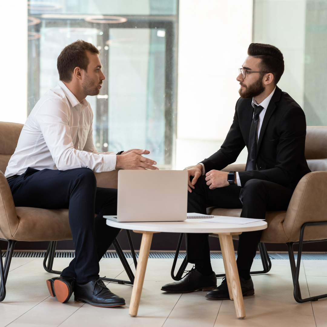 Two men in business attire having a meeting in a modern office, sitting on couches across from each other with a small white table and a laptop between them.