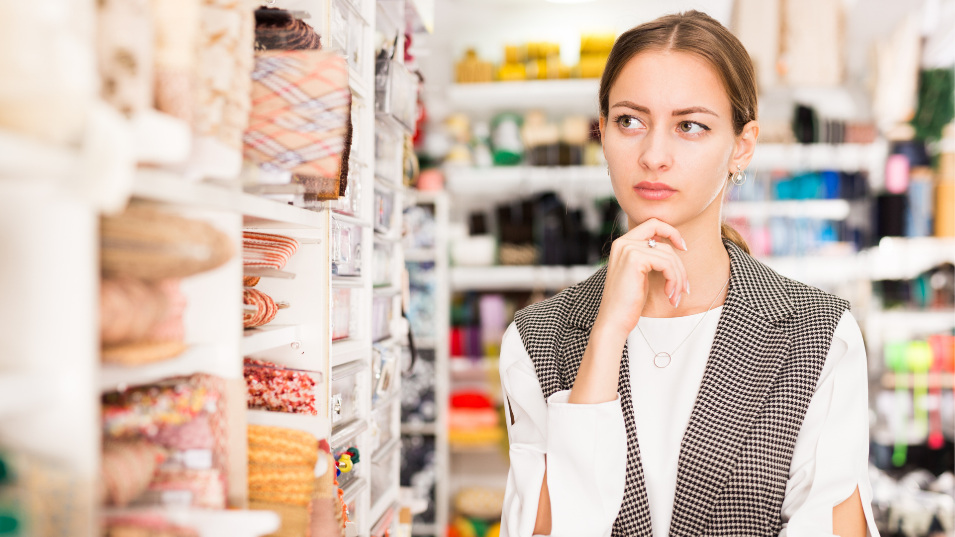 Woman shopping for fabric in a craft store, looking thoughtful as she examines the shelves of colorful fabric rolls.