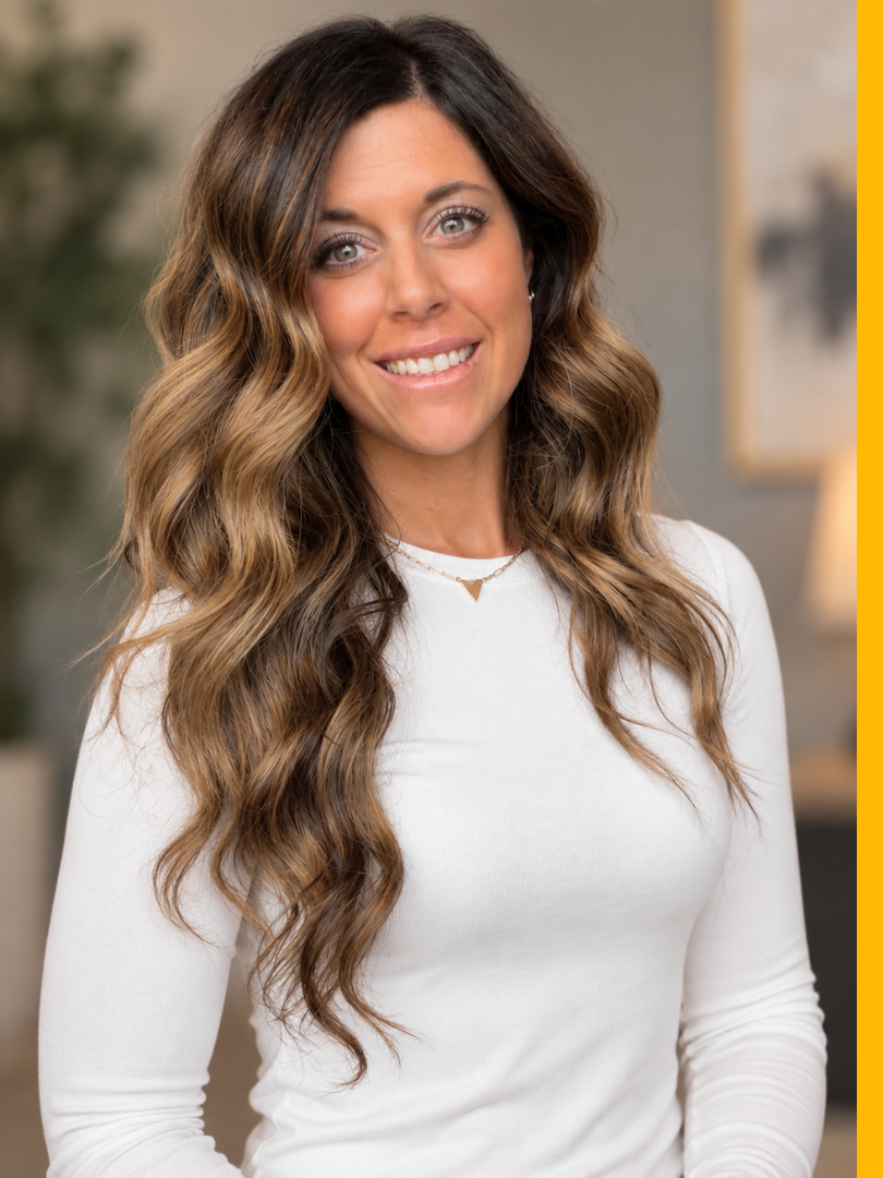 A young woman with long, wavy brown hair, wearing a white top and a gold necklace, smiling indoors with blurred background.