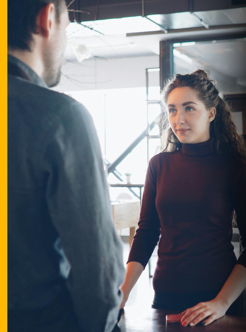 A young woman with curly hair and a maroon turtleneck engaging in a conversation with a man in a grey jacket inside a modern, well-lit space, possibly a cafe or office.