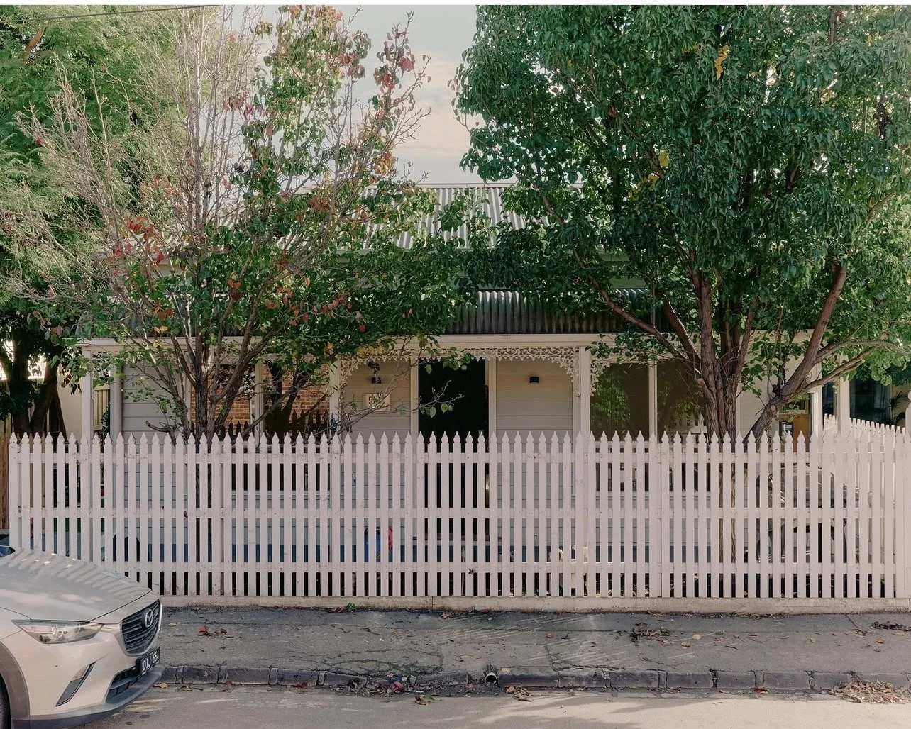 A house with decorative white picket fence and two large trees in front, with a silver car parked nearby.