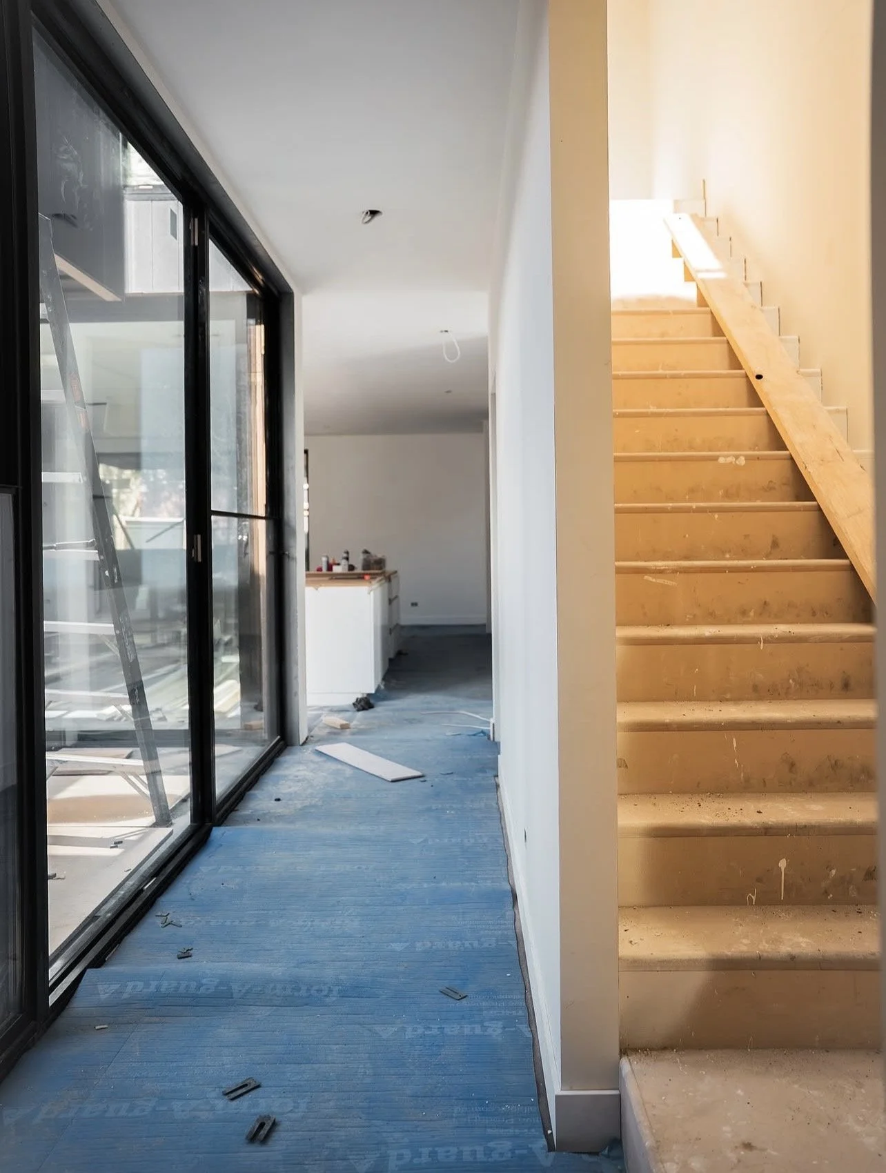 Interior of a house under construction with a partially finished staircase on the right, a glass sliding door on the left, and construction tools and materials scattered on the floor.