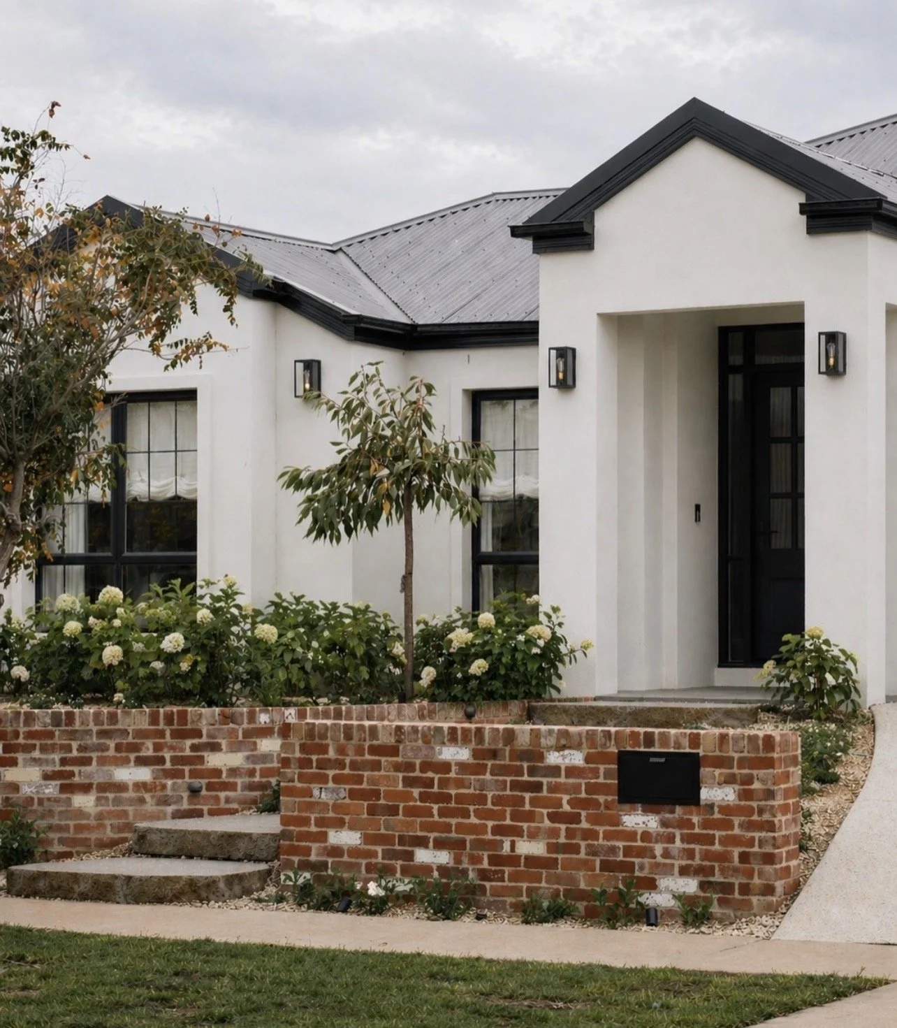 Modern white house with black roof and trim, surrounded by landscaped bushes and small trees, brick retaining wall in front, and steps leading to front door.