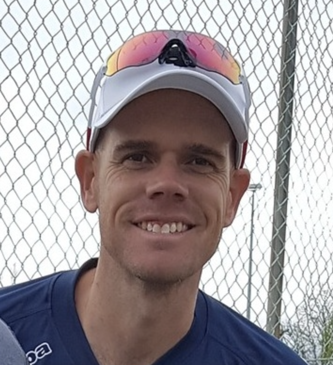Close-up of a smiling man wearing athletic gear, white cap with colored sunglasses on top, standing in front of a chain-link fence.