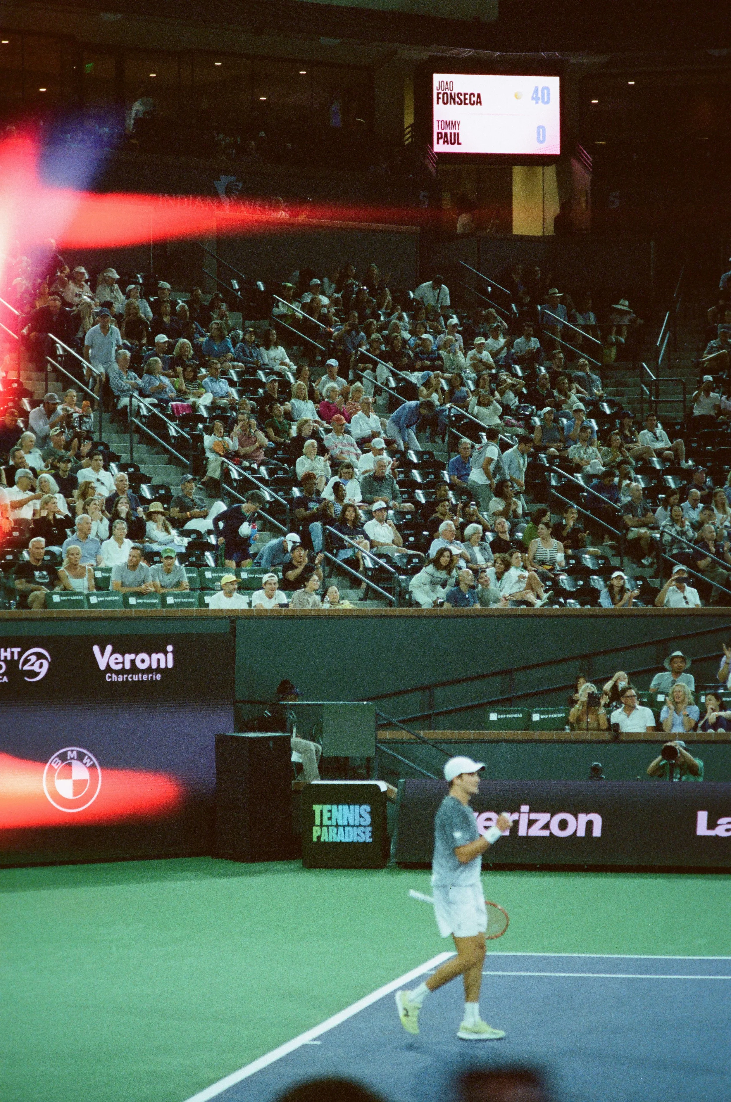 A tennis player on the court with an audience in the stands and banners displaying sponsors around the arena. The scoreboard shows Joao Fonseca leading Tommy Paul.