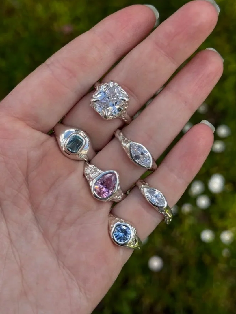 A hand displaying multiple rings with various large, colorful gemstones set in silver bands, outdoors with a blurred green background.