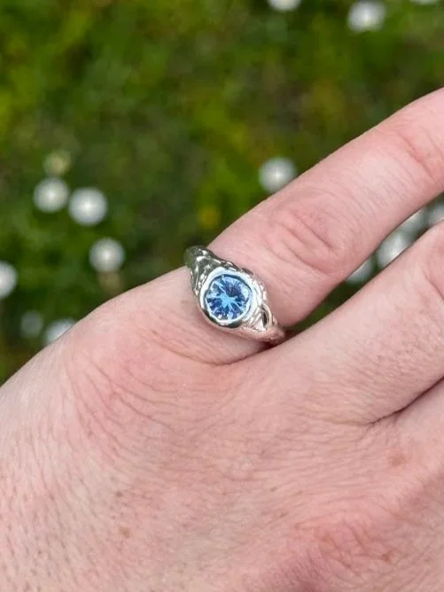 Close-up of a person's finger wearing a silver ring with a blue gemstone. The background is blurred greenery with small white berries.
