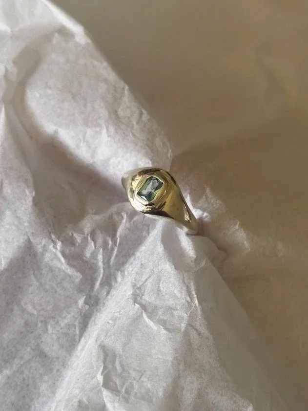 A gold ring with a rectangular dark gemstone, placed on crumpled white tissue paper.
