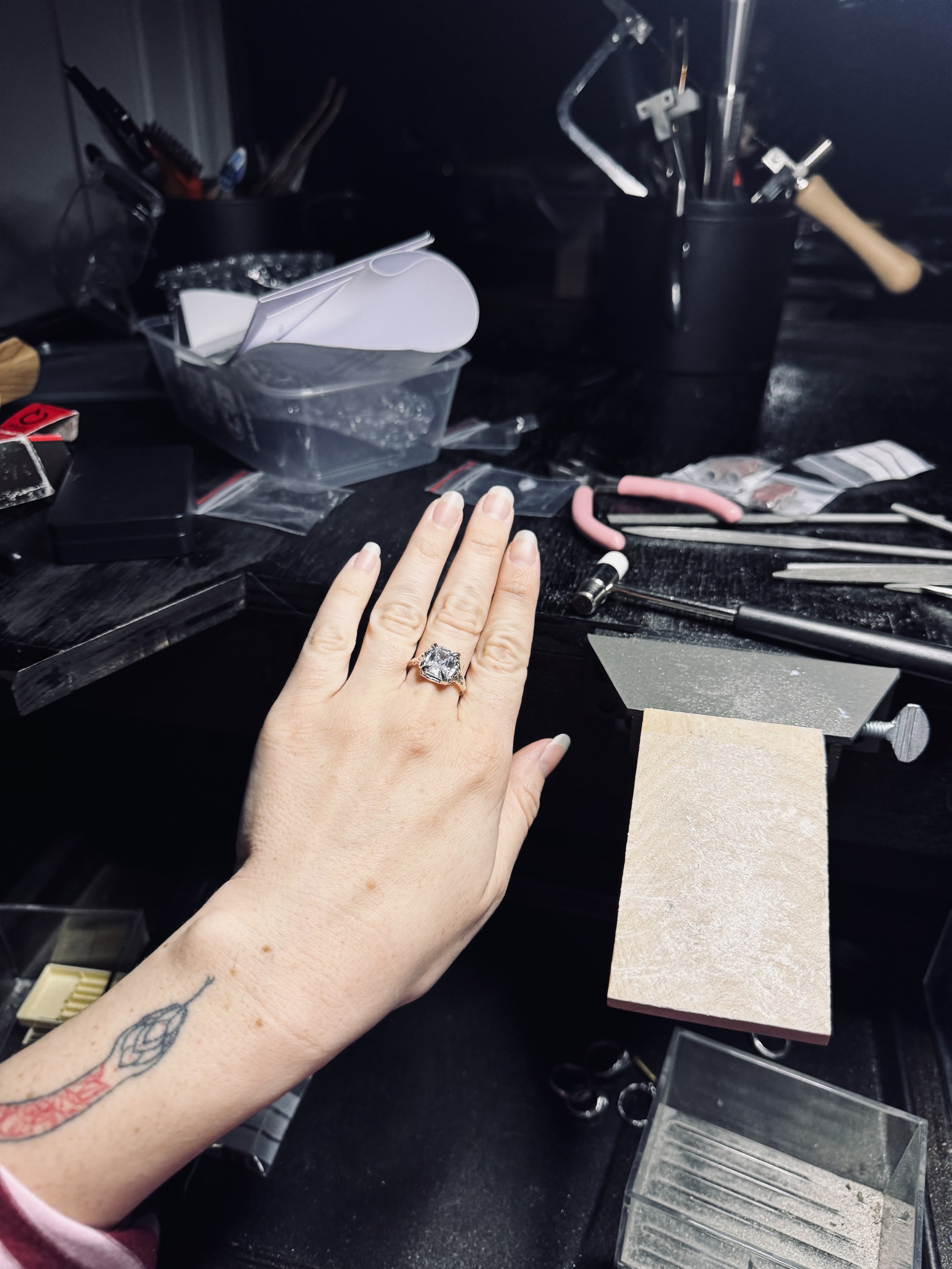 A person's hand with a decorative ring, tattoo, and light-colored nail polish resting on a black workbench surrounded by jewelry-making tools and supplies, including pliers, a sanding block, and small plastic containers for organizing jewelry components.