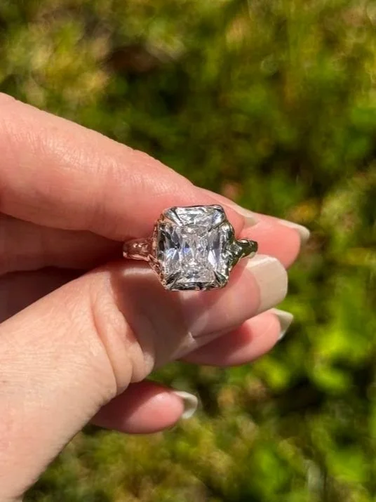 Close-up of a hand holding a large diamond ring with a rectangular stone, outdoors with green foliage in the background.