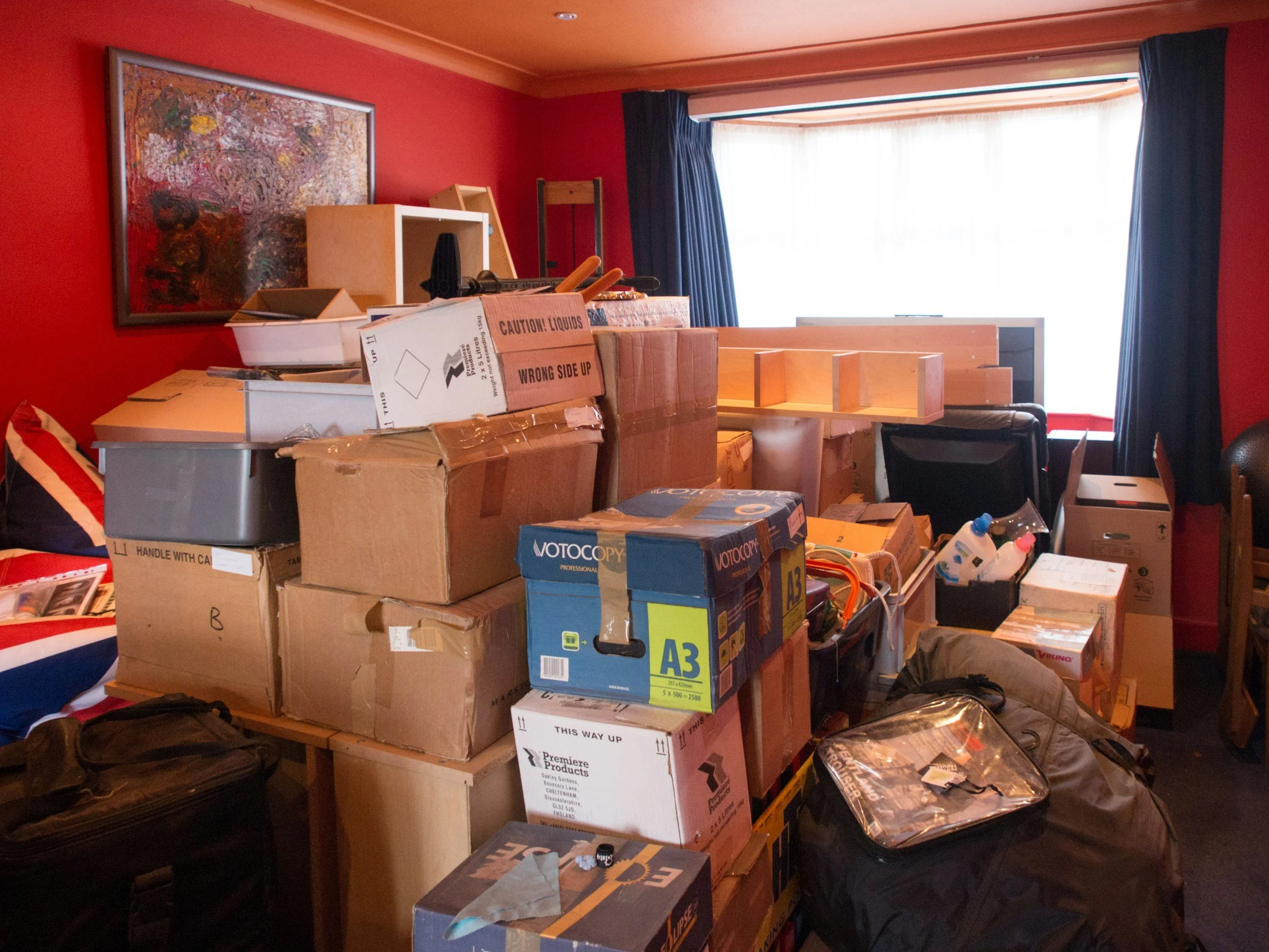 Cluttered red-walled room filled with stacked moving boxes, luggage, furniture, and household items.