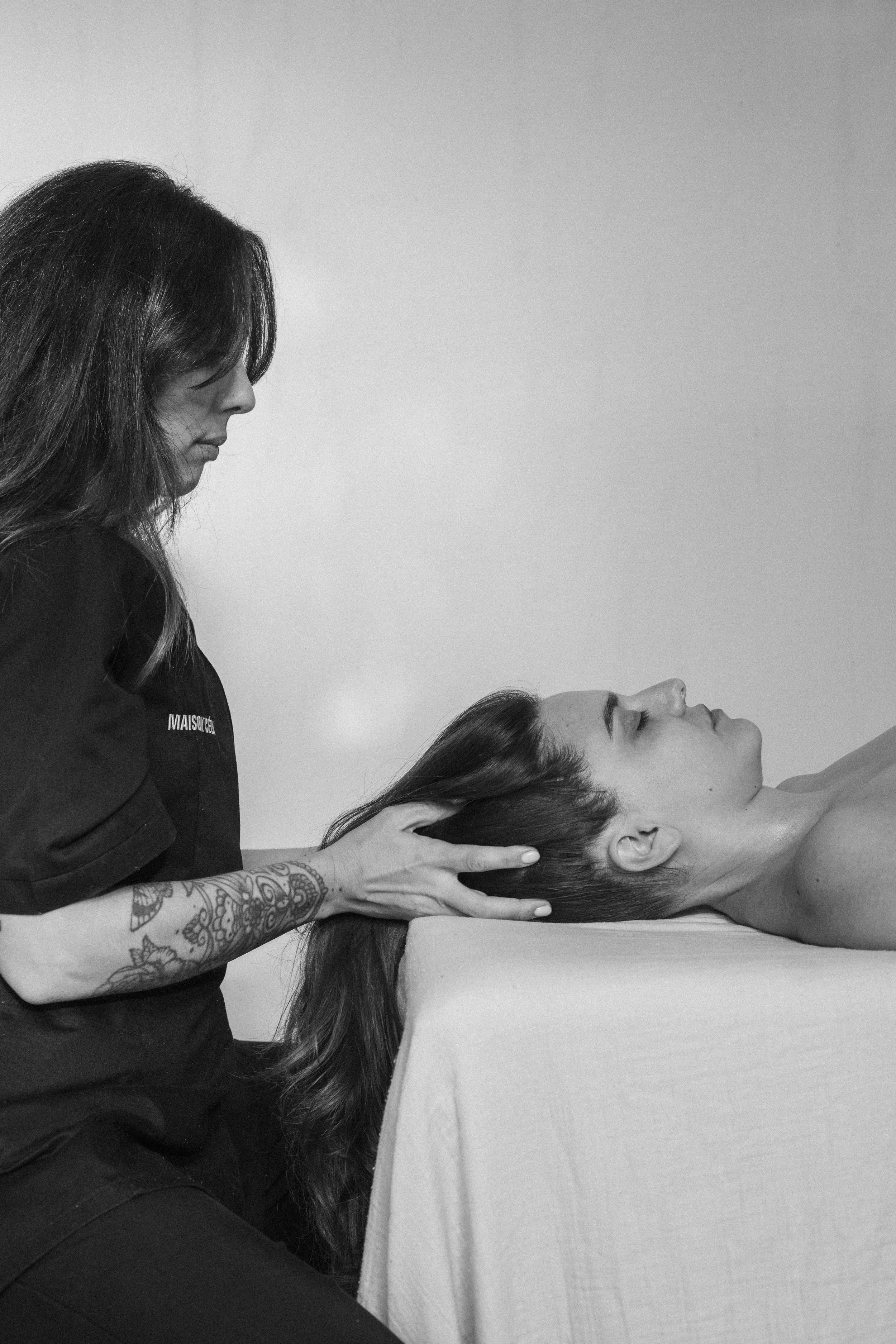Une femme reçoit un massage du cou et de la tête par une esthéticienne dans un environnement de spa, avec un fond lisse et clair.