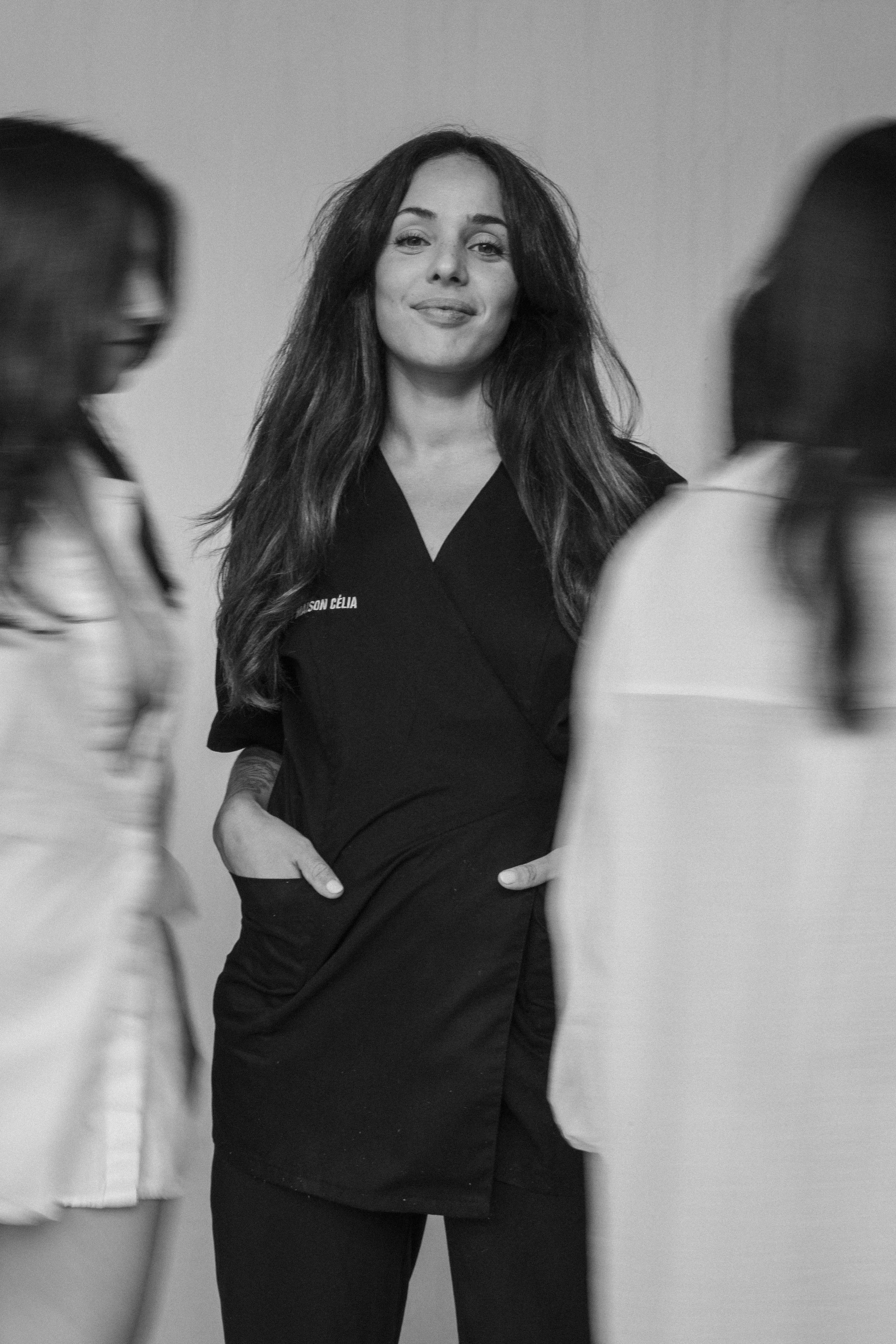 Une femme souriante avec des cheveux longs et ondulés, portant un vêtement noir, entourée de deux autres femmes floues dans un fond neutre.