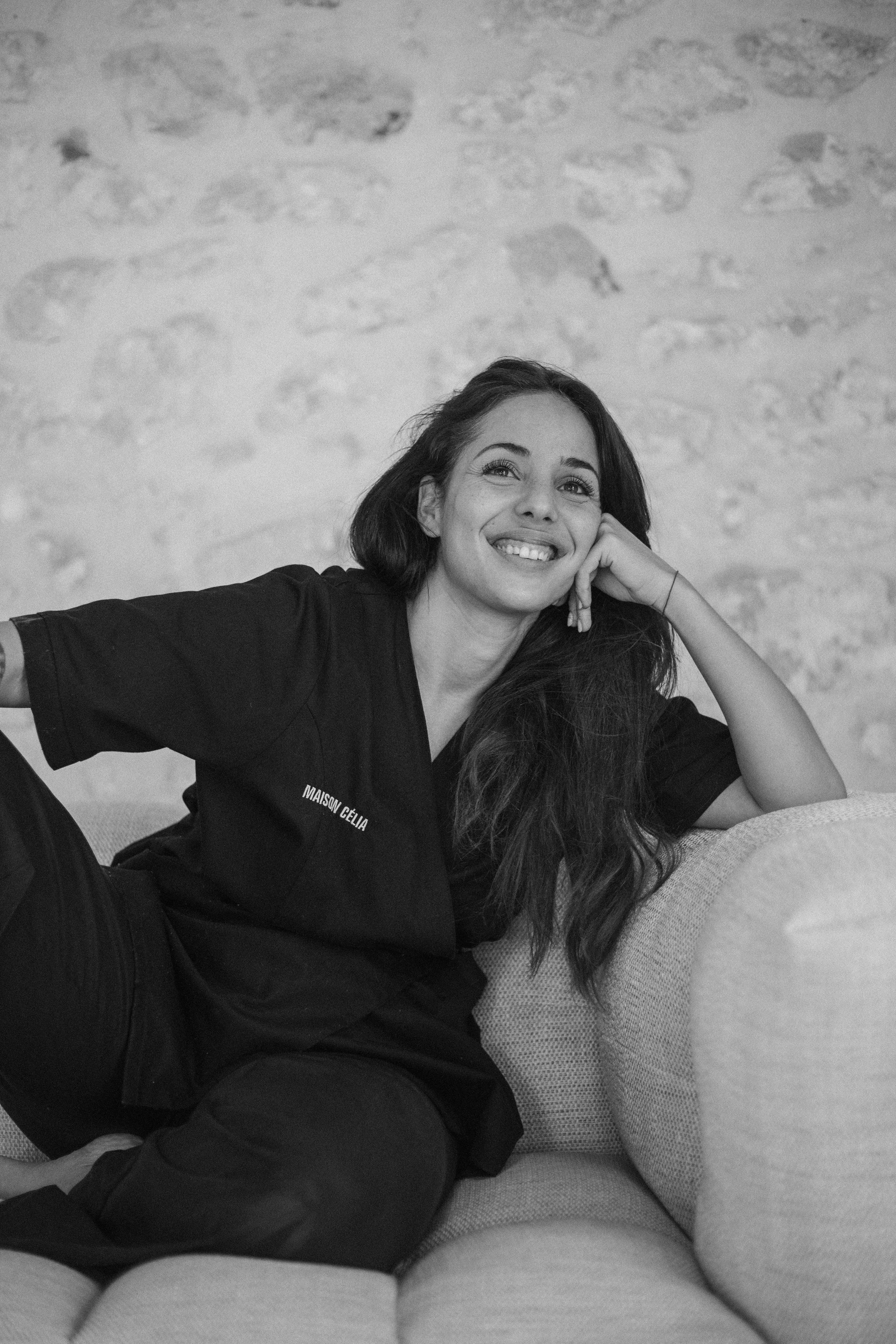 Une femme souriante repose sa tête sur la main, portant un t-shirt noir avec le texte 'Maison Celia', assise ou allongée sur un canapé ou un lit, en arrière-plan un mur en pierre.