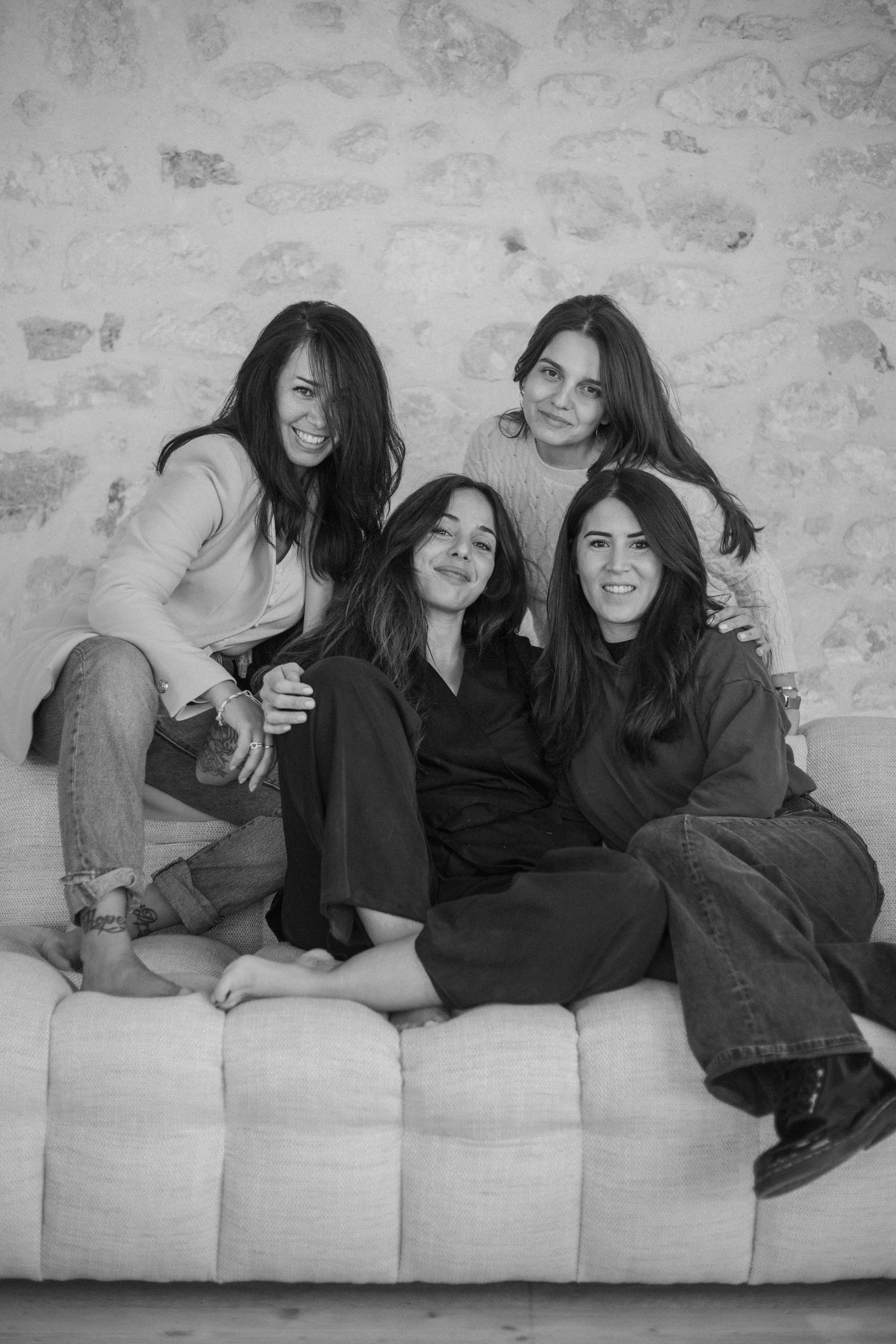 Quatre femmes jeunes posant ensemble sur un canapé, face à la caméra, en noir et blanc, dans un décor intérieur avec un mur en pierre en arrière-plan.