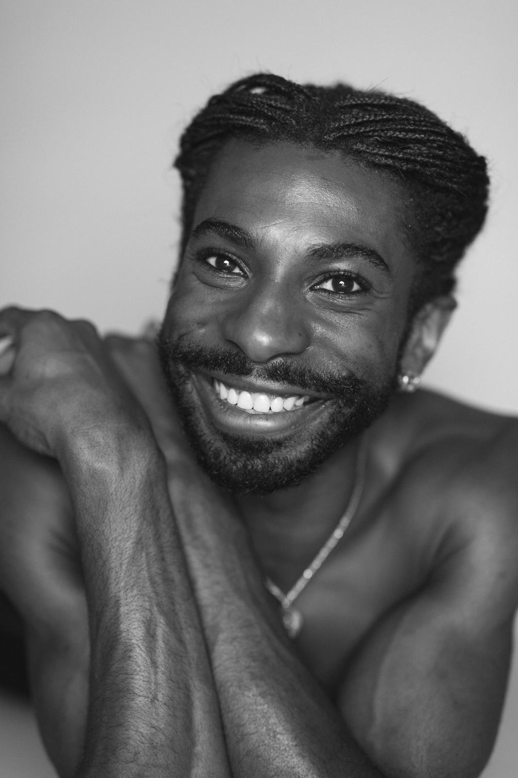Black-and-white close-up portrait of Emmanuel Diela Nkita displaying braided hair, beard, and necklace.