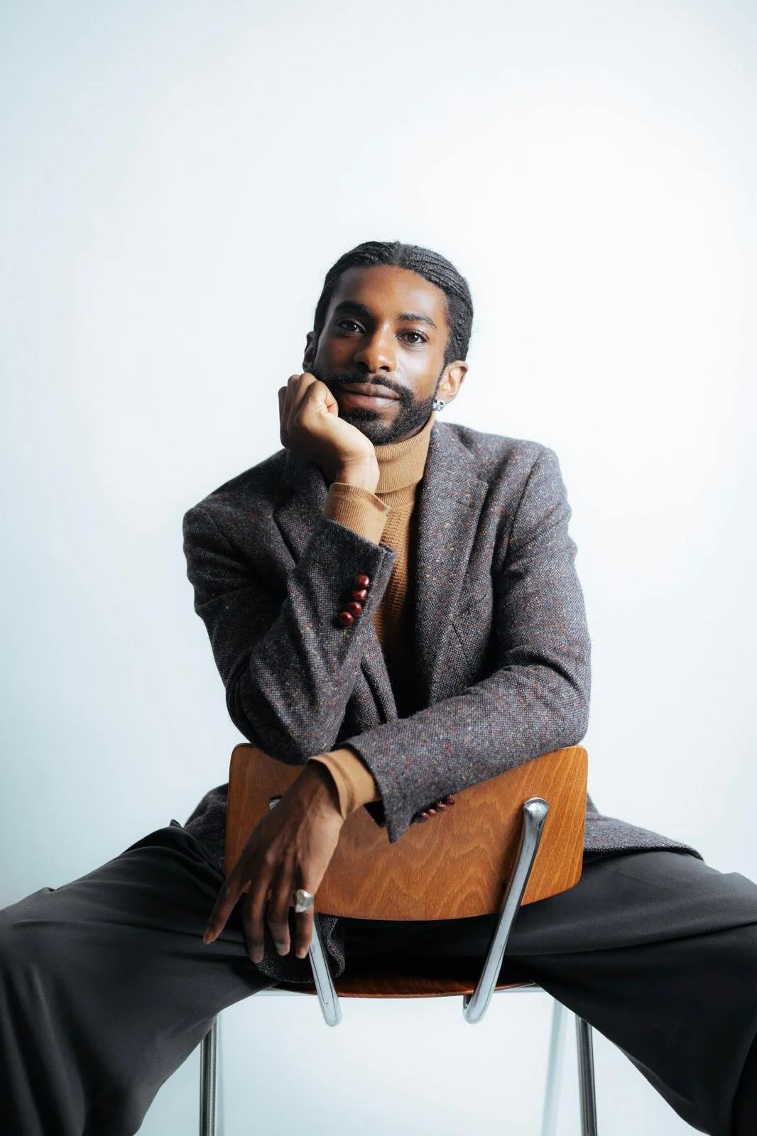 Portrait of Emmanuel Diela Nkita sitting backward on a wooden chair, wearing a blazer and turtleneck sweater.