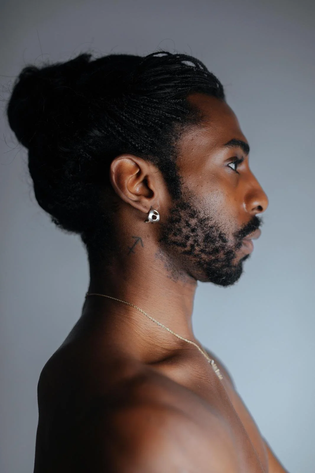 Side-profile portrait of Emmanuel Diela Nkita displaying braided hair in a bun, beard, earrings, and necklace