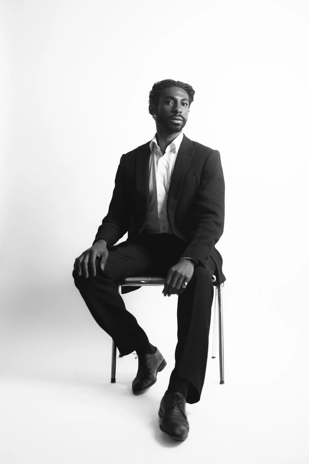 Black-and-white portrait of Emmanuel Diela Nkita in a suit sitting on a metal chair against a white background