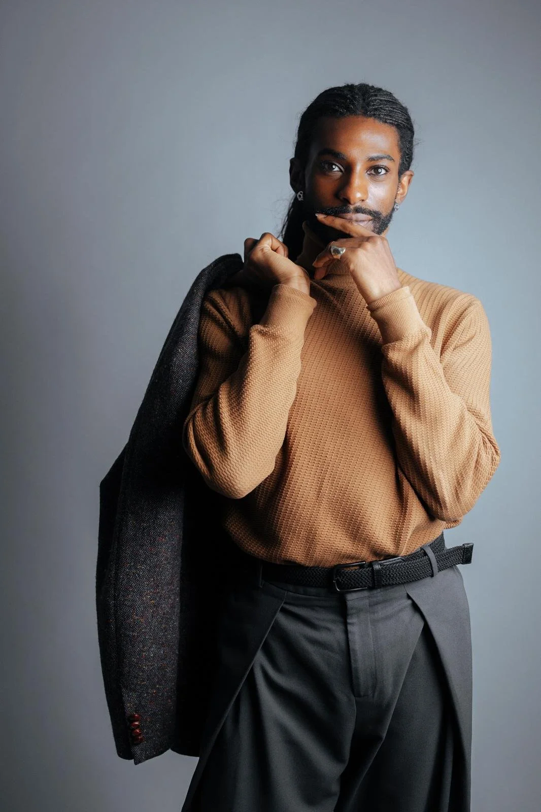 Fashion portrait of Emmanuel Diela Nkita wearing a tan sweater, black trousers, and a draped dark coat
