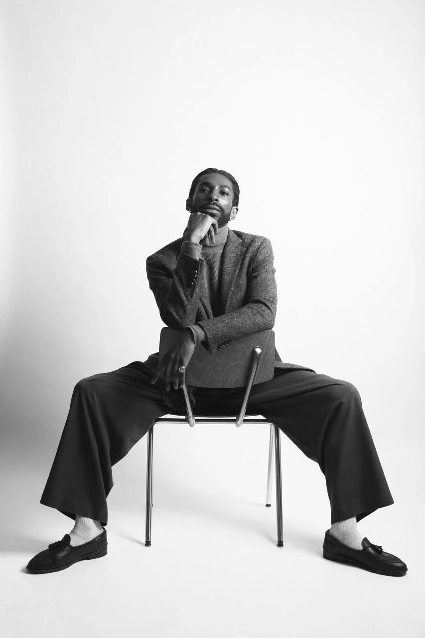 Black-and-white portrait of Emmanuel Diela Nkita sitting backward on a chair with his chin resting on his hand.