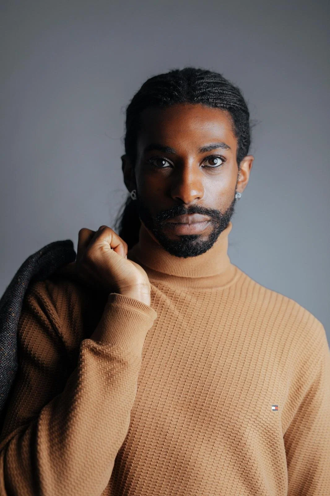 Portrait of Emmanuel Diela Nkita wearing a tan textured turtleneck sweater against a gray background.