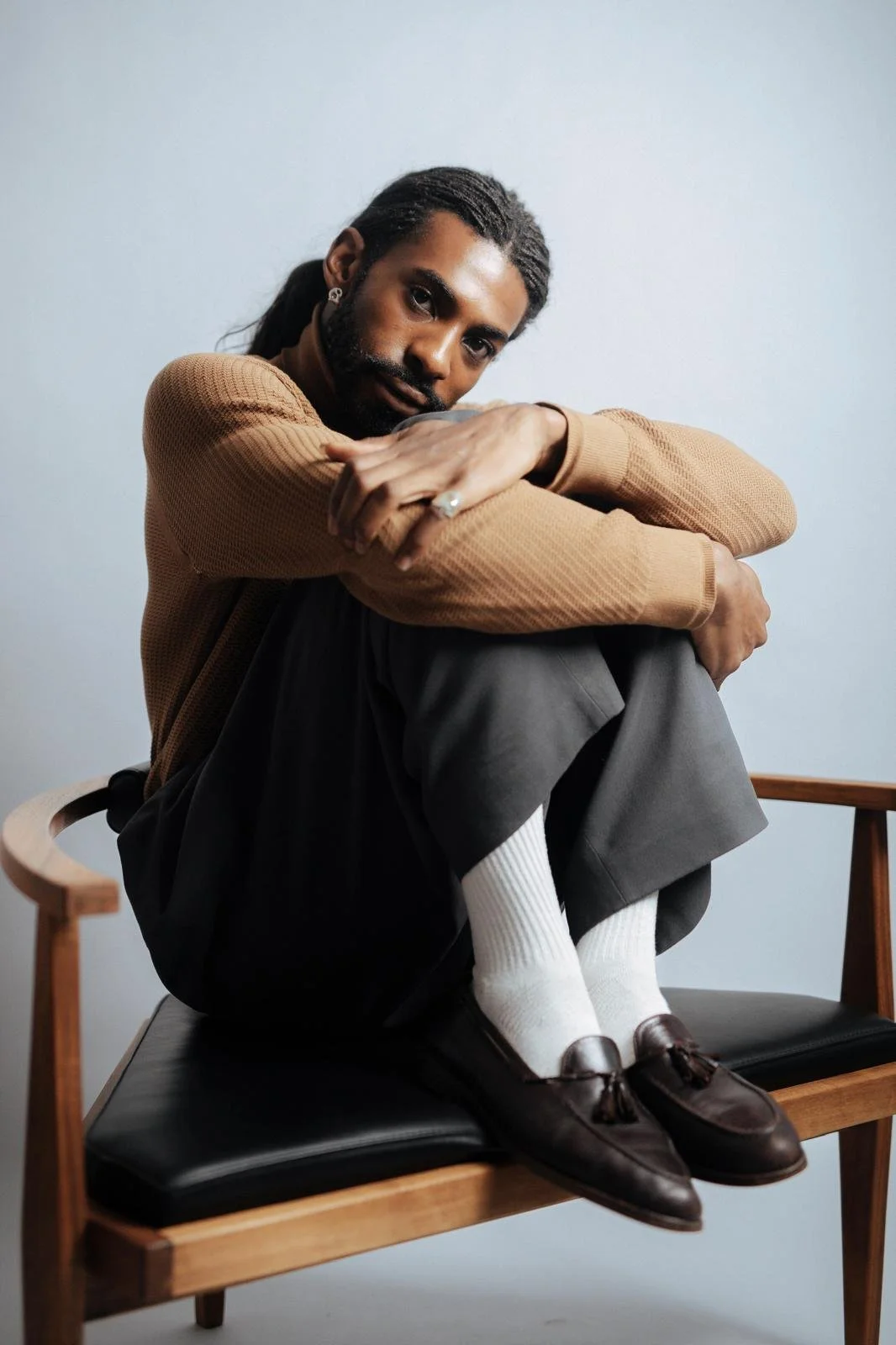 Portrait of Emmanuel Diela Nkita sitting cross-legged in a wooden chair, wearing a brown sweater, gray trousers, white socks, and brown loafers.
