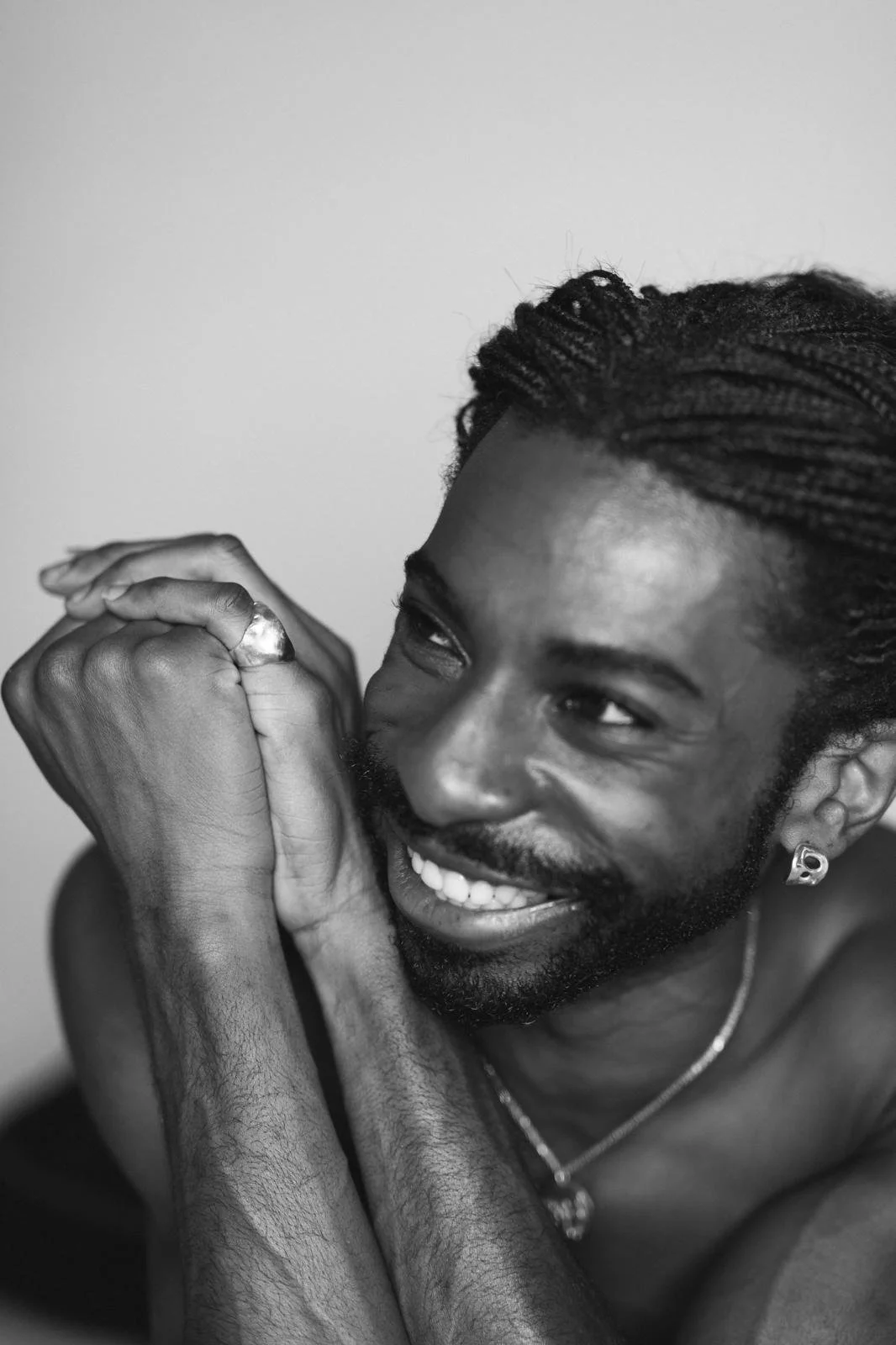 Black-and-white close-up portrait of a smiling bearded man with braided hair, earrings, a ring, and necklace.