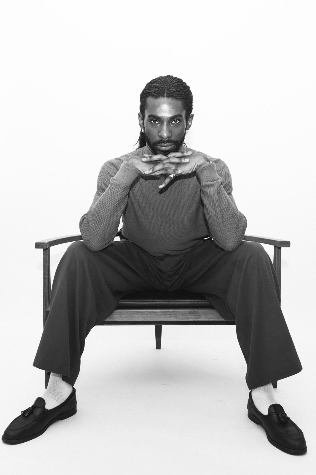 Black-and-white portrait of Emmanuel Diela Nkita sitting in a wooden armchair, wearing a textured sweater, wide-leg pants, socks, and loafers.
