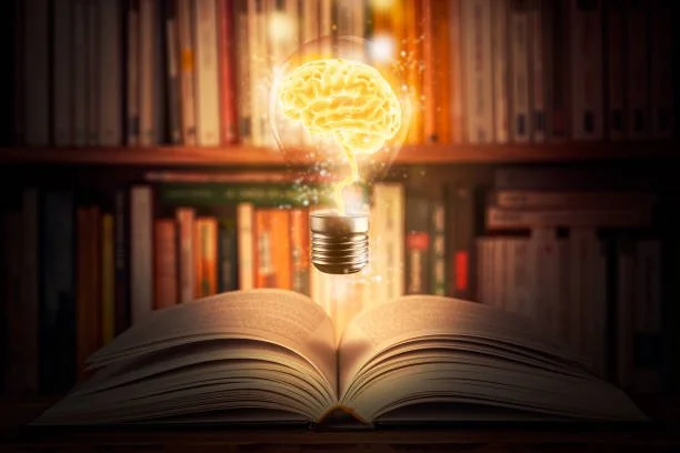 An open book on a wooden surface with a glowing light bulb and illuminated brain hologram above it, with a blurred bookshelf background.