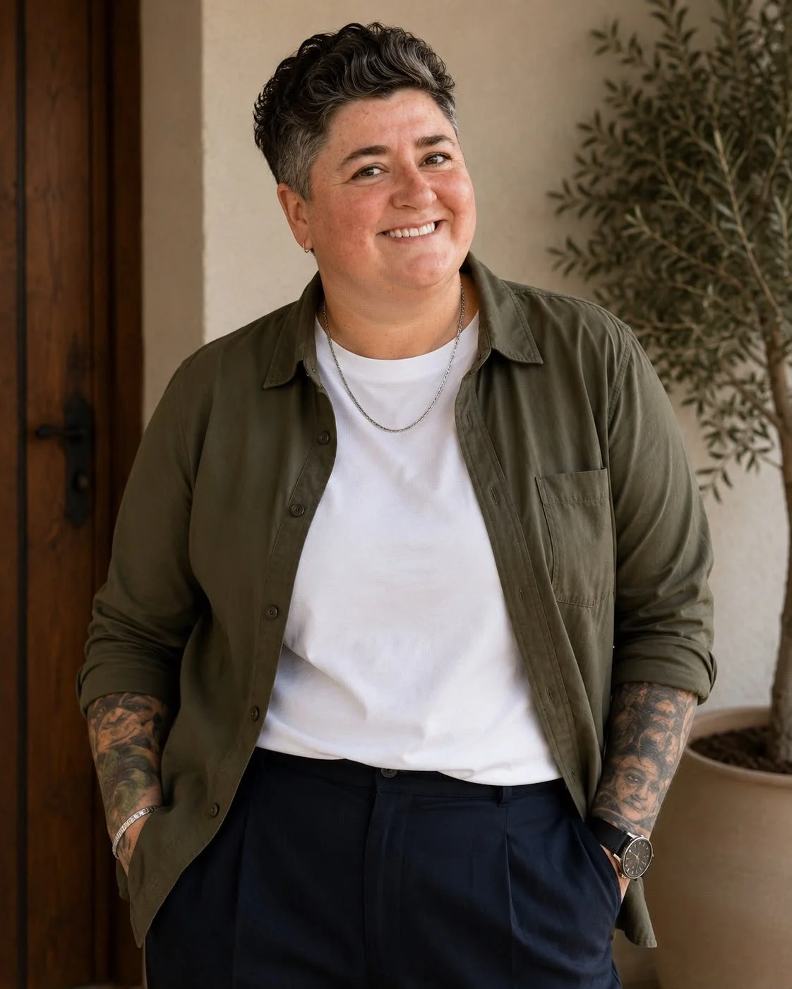 A smiling woman with short curly hair, wearing a white T-shirt, an olive green button-up shirt, dark pants, and accessories, standing indoors with a potted plant in the background.