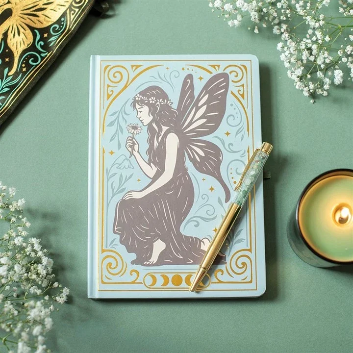 A hardcover book featuring a fairy illustration on the cover, with fairy wings, sitting and holding a flower. The book is surrounded by white flowers, a lit candle, and a gold and clear pen, on a pastel green surface.