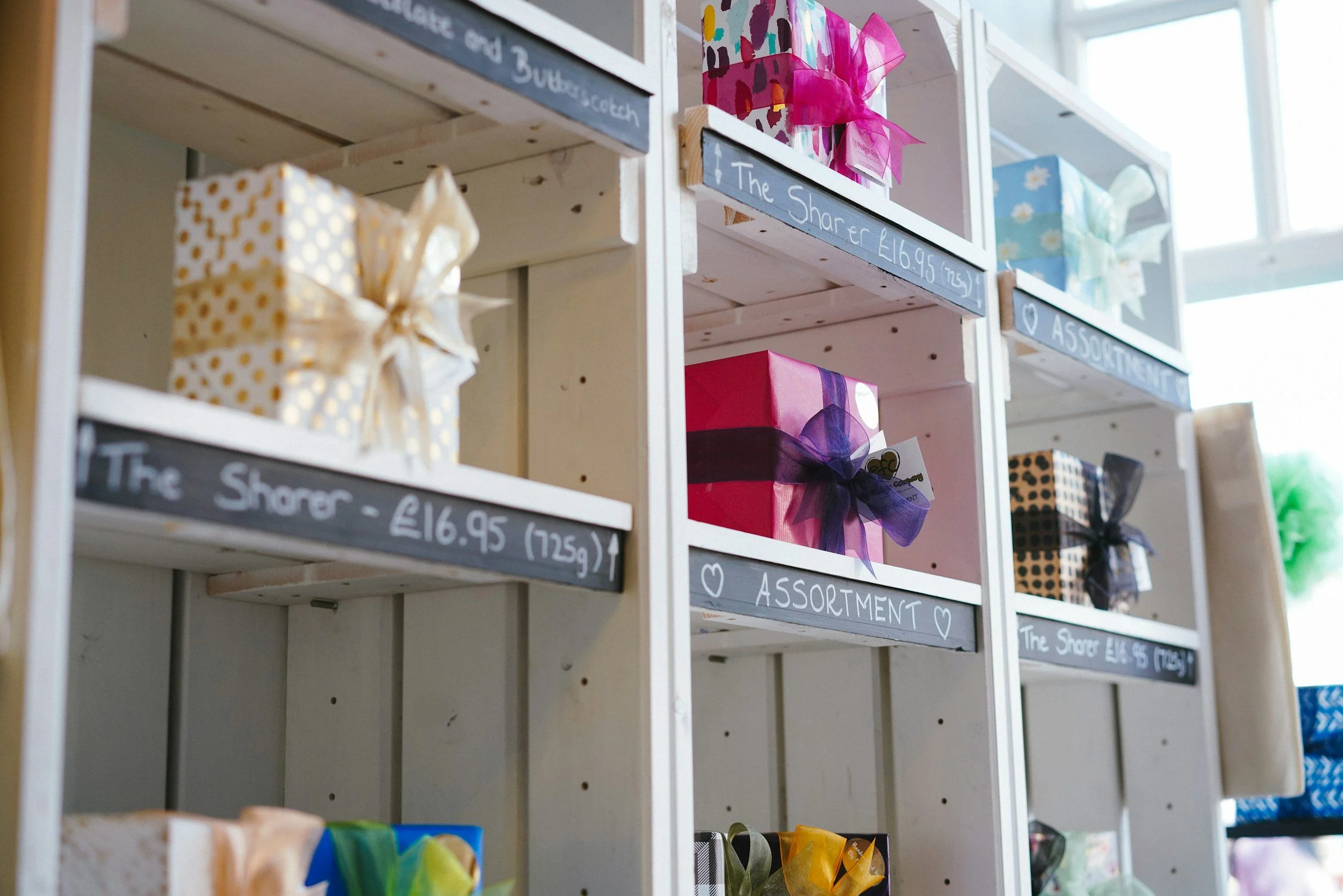 Wooden shelves with gift boxes wrapped in colorful paper and ribbons, labeled with handwritten tags such as 'The Sharer' and 'Assorted', inside a store.