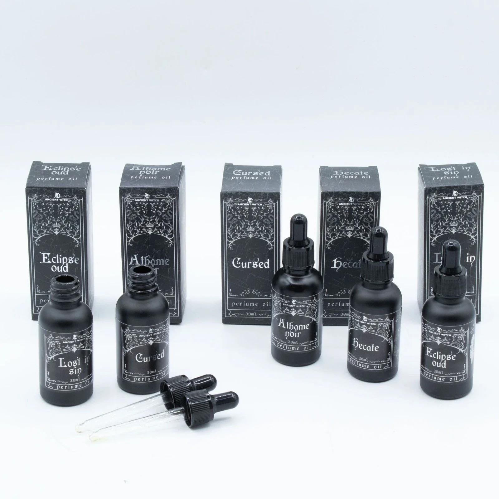 Set of five black bottles labeled with different perfume names, including 'Eclipse Oud,' 'Alfame Noir,' 'Cursed,' 'Hecate,' and 'Lost in Sin.' Some bottles have dropper caps, and there are black boxed labels behind each bottle matching the perfume names.