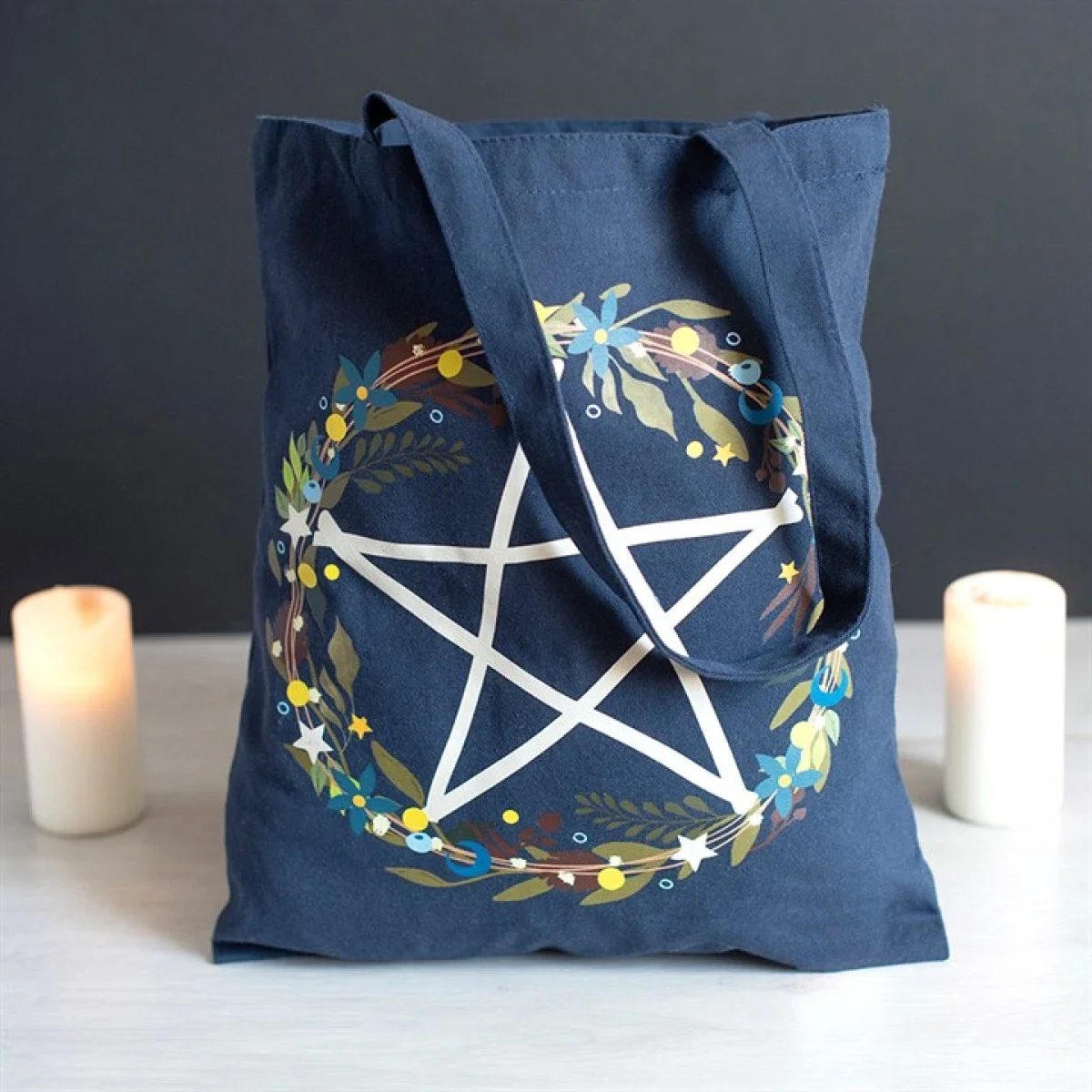 A blue tote bag decorated with a white pentagram and a floral wreath design, flanked by two candles on a white surface against a dark background.