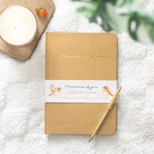 A closed journal with a tan cover and gold writing that says "Memorize of you," placed on a white textured surface. Next to the journal is a gold pen, and above it, a round wooden tray holding a small candle and a small decorative object. There is a blurred green plant in the background.