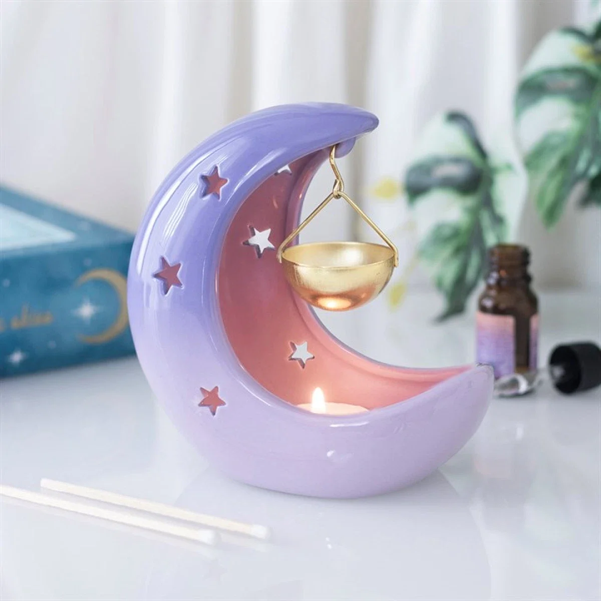 Decorative moon-shaped lamp with stars, a hanging gold bowl, and a lit candle inside the moon, placed on a white surface.