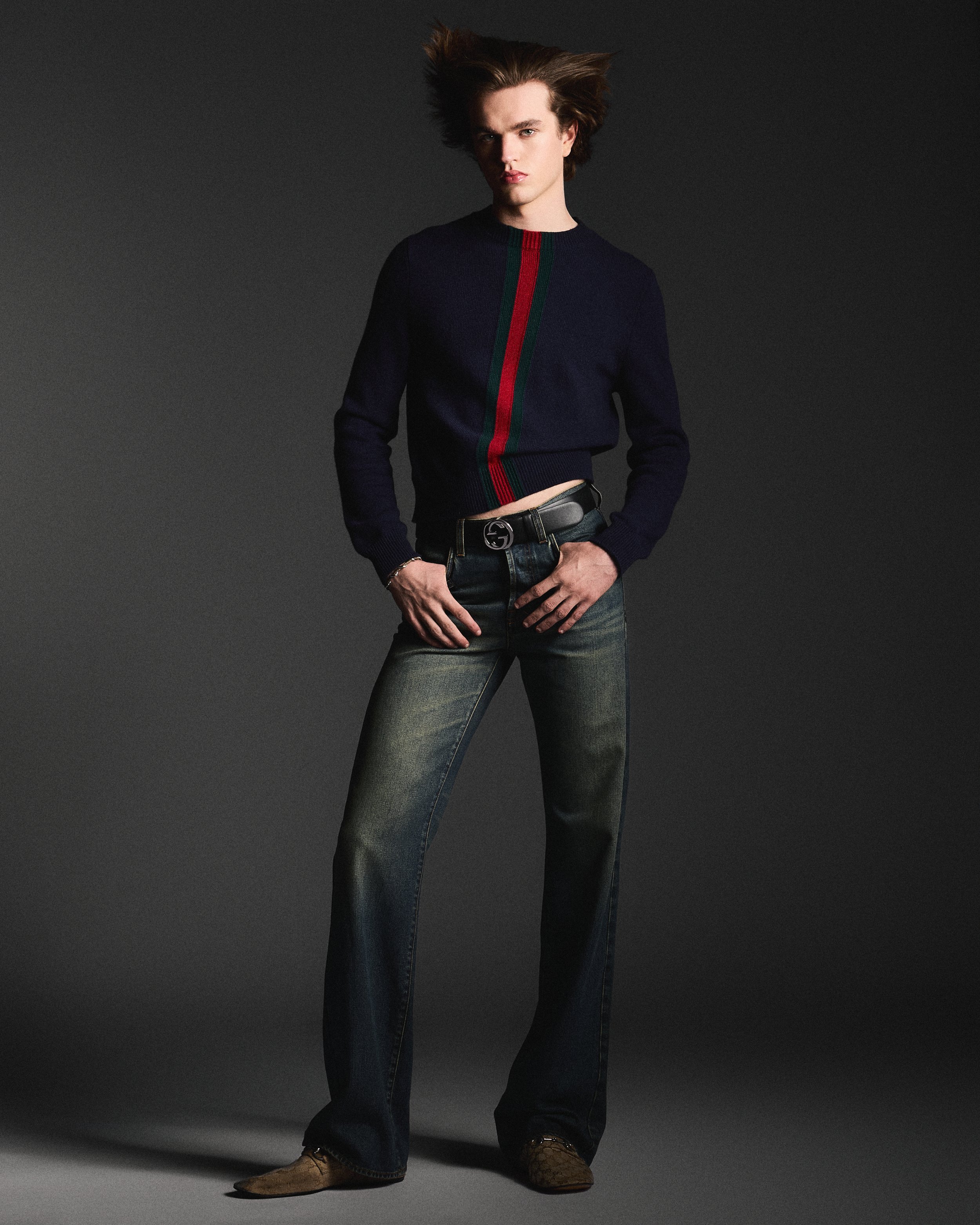 A young man with styled hair wearing a dark navy sweater with red and green vertical stripes, blue jeans, brown cowboy boots, and a belt with a distinctive buckle, standing against a dark background.