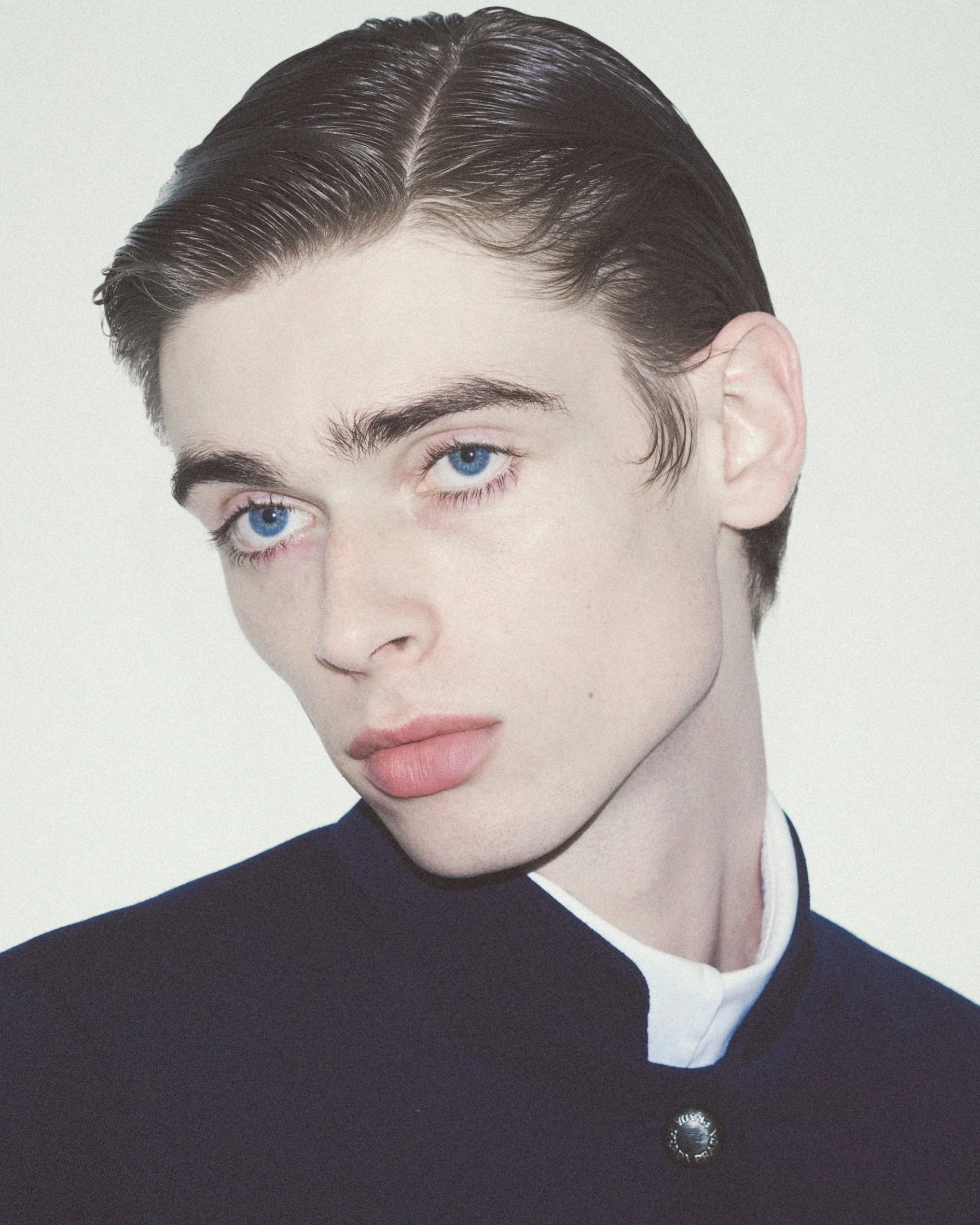 Close-up of a person with dark, slicked-back hair, fair skin, and bright blue eyes, wearing a black top with a white collar.