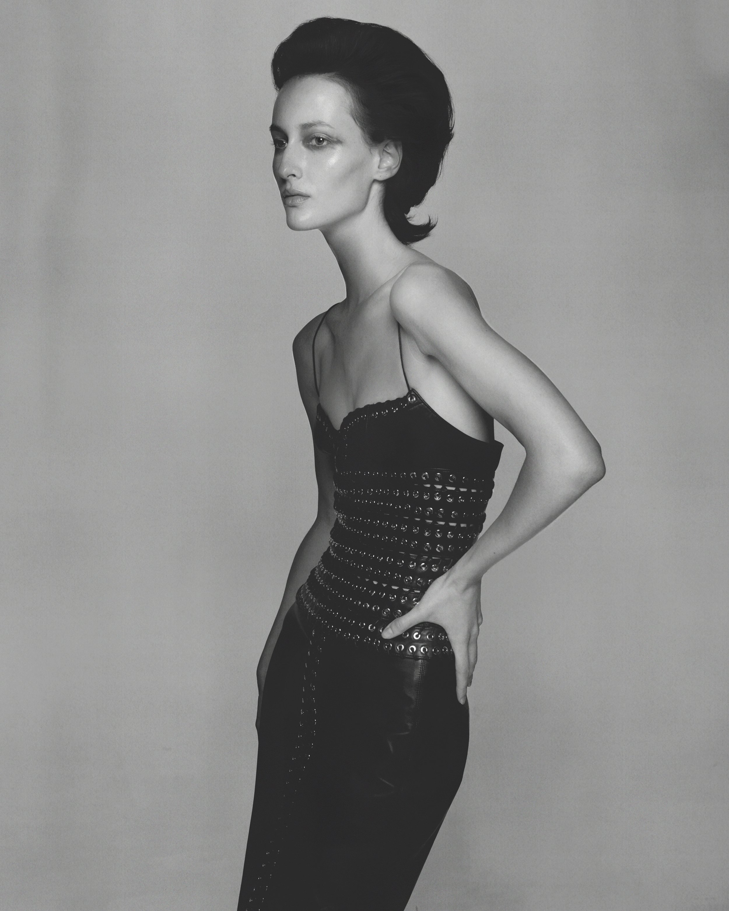 Black and white photo of a woman with short, dark hair styled back, wearing a black, strapless dress with embellishments, standing in profile against a plain background.