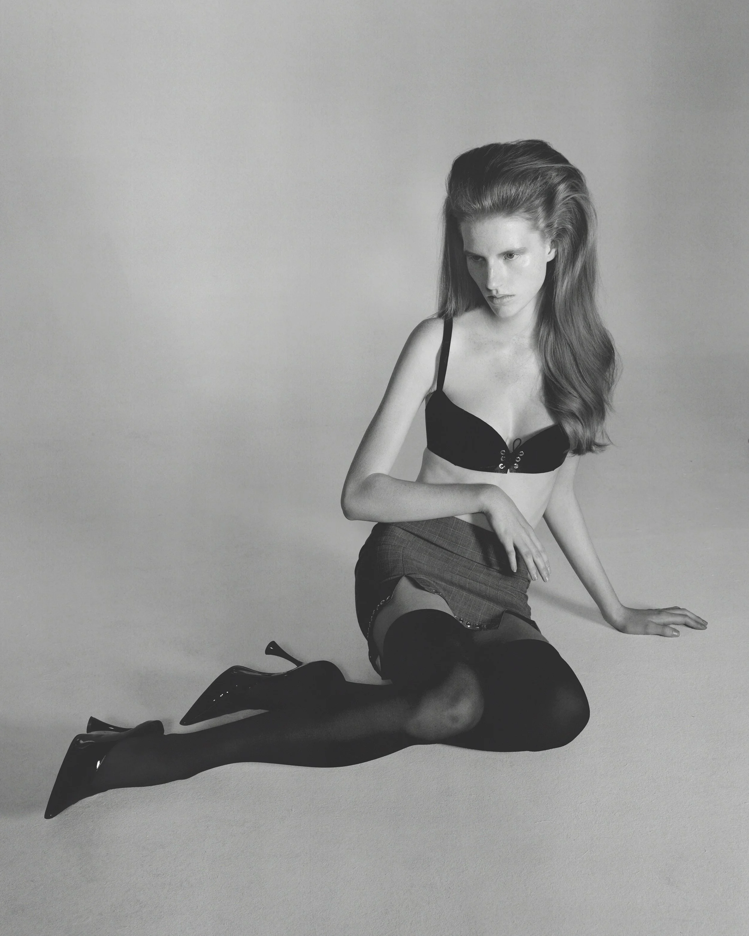 A black-and-white photograph of a woman with long hair, wearing a black bra, sitting on the floor with her legs bent and her hands resting on her knee and the floor. She is wearing high-heeled shoes and a skirt. The background is plain and light-colored.
